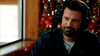 A bearded man in a green plaid shirt and headphones sits by a window, in front of a decorated Christmas tree with red ornaments and lights, ready to watch the "Christmas on the Ranch (2024) DVD.