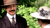 A mustached man in a brown suit and straw hat chats with a woman in a white hat with silver ribbon outdoors, as they discuss "The Brides in the Bath (2003) DVD," a true crime story about George Joseph Smith.