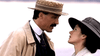 A man in a straw boater and overcoat speaks to a woman in a wide-brimmed hat and light coat—an evocative moment recalling "The Brides in the Bath (2003) DVD," set outdoors against a softly blurred background.