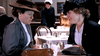 Two women in early 1900s attire sit at a tea room table, chatting about The Brides in the Bath (2003) DVD as teapots and a purse rest before them, while a waitress serves customers in the background.