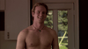 A shirtless young man with light brown hair smiles indoors near a glass door, trees outside. He wears a black necklace with a small pendant, ready to watch "A Friend of the Family (2005)" on DVD—a gripping thriller.