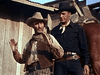 Two men dressed as cowboys stand outside a wooden building—one in a tan hat and vest with hands raised, the other in black looking serious—just like scenes from Carson City (1952) DVD.
