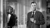 Two men indoors: one in a suit holding a cigarette, looking serious; the other, a friendly butler peeking through a door—a scene reminiscent of classic films like Queen Bee (1955) DVD.