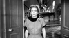 A woman with short, styled hair and bold brows stands in a doorway, evoking Joan Crawford in Queen Bee. She wears a dark fur-trimmed dress with a checkered floor behind her. Experience classic film drama with Queen Bee (1955) on DVD.
