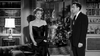 A woman in a chic black dress and pearls stands by a suited man in a refined library, channeling Joan Crawford’s elegance—ideal for classic film enthusiasts seeking the Queen Bee (1955) DVD.