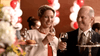 A well-dressed woman and man smile and clink wine glasses at a festive event, with balloons in the background and trays of pastries, flowers, and the "An Officer and a Murderer (2012) DVD" displayed in the foreground.