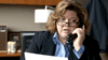 A woman with shoulder-length brown hair and glasses, in a dark blazer and blue shirt, sits at a desk holding a phone and looking at a document. Office supplies, photos, and the "An Officer and a Murderer (2012) DVD" are in the background.