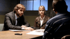 A man in a suit and a woman in a brown leather jacket sit at a table with papers, facing someone whose back is to the camera—an intense scene from the "An Officer and a Murderer (2012)" DVD.