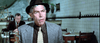 A man in a gray suit, brown shirt, patterned tie, and dark fedora sits at a restaurant table, capturing the vintage style seen in the Hard Times (1975) DVD. Another man is in the background amid white tiled walls.