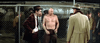 A shirtless man stands near a chain-link fence at night, tense and ready to fight, surrounded by men in suits—just like scenes from "Hard Times (1975)" on DVD.