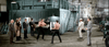 Two shirtless men box in a warehouse while suited men and one man in white watch—some on the floor, others above. This iconic scene from "Hard Times (1975)" is available on DVD.