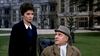 A woman in dark clothing stands behind an older man in a coat and hat, seated outdoors by a grand historic building—a scene recalling the suspense of Woman of Straw (1964) DVD.