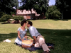 A man and woman enjoy a sunny park picnic, with the man leaning in to kiss her cheek. In the background stands a large house reminiscent of settings from "A Woman Named Jackie (1991) DVD.