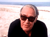 An older man with gray hair in black sunglasses and a dark shirt sits on a sandy beach by the ocean, enjoying the DVD "A Woman Named Jackie (1991).