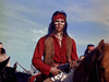 A person with long dark hair, wearing a red shirt, brown vest, and red headband with white face paint, sits on horseback holding a pistol—evoking vintage scenes from classic westerns like the Fort Yuma (1955) DVD. Blue sky and riders are in the background.