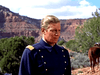 A man in a blue military uniform with gold buttons stands outdoors in a rocky desert with shrubs and red cliffs, evoking classic scenes from Fort Yuma (1955) DVD. A horse is partially visible behind him.