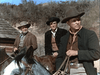 Three men in cowboy hats and vintage Western clothes are outdoors; two are on horseback, one stands nearby. A wooden building and dry hills set the scene—ideal for fans of rare films like Five Guns West (1955) DVD.