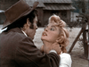 A man in a brown hat grabs a blonde woman by the neck and stares at her intensely outdoors near a rustic wooden building—a gripping scene reminiscent of rare films like Five Guns West (1955) DVD. The woman looks up at him, tense.