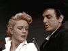 A worried blonde woman in a white blouse looks away as a serious man in a black hat and jacket stands beside her with a gun pointed up—a dramatic scene from rare films like Five Guns West (1955) DVD.