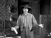 A man in a wide-brimmed hat, striped shirt, and vest stands by a brick wall and wooden door, gesturing with both hands. A chair and hanging gourds are beside him. Black-and-white still from The Broken Star (1956) DVD.