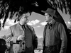 Two men converse outdoors under a tree—one in a suit with a glass, the other wearing a cowboy hat and vest. Bottles sit on the table with mountains behind them. Scene from "The Broken Star (1956)" DVD.