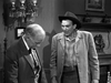 Two men in vintage Western attire stand indoors; one looks down while the other, in a cowboy hat and coat, watches him. A chandelier, framed art, and a fireplace evoke the classic charm of "Five Guns to Tombstone (1960) DVD.