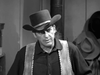 A man in a cowboy hat and vest stands indoors, glancing sideways. Several hats hang on the wall and a stove pipe appears behind him in this black-and-white scene from "Five Guns to Tombstone (1960)" DVD.
