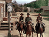 Three men in cowboy hats ride horses down a dusty street past a large clock and wooden building with steps—just like a scene from The Cimarron Kid (1952) DVD.