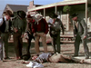 Six cowboys in western gear surround a man lying on the ground in front of a rustic building, capturing the suspenseful atmosphere of "The Cimarron Kid (1952)" DVD, set in a dusty old western town.
