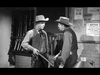 Two men in cowboy hats and western attire talk in a rifle- and poster-lined room. One holds a shotgun, the other leans on a chair. This black-and-white scene evokes classic Westerns like Gunfighters of Abilene (1960) DVD.