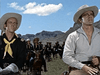 Two men in cowboy hats and bandanas stand in the foreground of a classic scene from "Comanche (1955) DVD," with riders on horseback and rocky mountains under a blue, cloud-filled sky in the background.