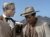 Two men evoke vintage Western style outdoors: one in a tan cowboy hat and rugged jacket, the other in a ruffled shirt and suit. Mountains and people are in the background. Featured on the "Comanche (1955) DVD.