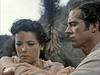 A concerned dark-haired woman examines her arm while a man with short dark hair watches. Both look worried, set against foliage and a blurred mountain—capturing the intensity of Comanche (1955) DVD.