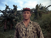 A man in a camouflage shirt and red cap stands outdoors by banana trees and lush green foliage under a cloudy sky, evoking scenes from vintage classics like the Ambush Bay (1966) DVD.