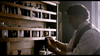 A man in a white shirt and dark vest examines a small brown bottle by shelves of jars in a dim room, creating a scene reminiscent of the historical drama in Effigy: Poison in the City (2019) DVD.