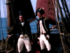 Two men in 18th-century naval uniforms stand on a ship with red sails, echoing scenes from the rare "Sea Devils (1953) DVD." They wear bicorne hats, dark coats with gold buttons, and white trousers against an ocean backdrop.