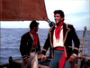 Two men in historical naval uniforms stand on a wooden sailboat at sea, one smiling and holding the ship’s wheel. Both wear dark jackets with gold trim, as seen in "Sea Devils (1953)"—available now on DVD. The ocean stretches out behind them.
