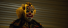 A figure in a creepy clown mask with red, white, and blue features, yellow hair, and a dark ruffled costume stands before corrugated metal—evoking scenes from the "Haunt (2019) DVD.