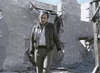 A man in a leather jacket navigates building ruins with a weapon in hand, capturing the tense atmosphere of Warhead (1977) DVD, set among debris and broken walls under a clear sky.