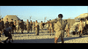 A group of soldiers in light uniforms stand on sandy ground near tents and a truck, barbed wire visible. This tense WWII scene is from Tank Force (1958) DVD—perfect for classic war movie fans. One man faces the group with arms outstretched.