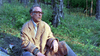 An older man in glasses and a beige coat sits thoughtfully on the grass in a sunlit forest, as if reminiscing about watching Citizen X (1995) on DVD.