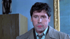 A man in a tan trench coat and striped shirt blinks and looks around uneasily before a blue wall—resembling a character straight out of Citizen X (1995) DVD.