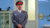 A man in a gray military uniform and red-trimmed hat stands indoors, evoking scenes from Citizen X (1995) DVD as he talks and blinks in front of a white double door and light blue walls.