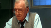 An older man in glasses, a blue shirt, tie, and gray sweater vest sits at a dimly lit table, looking down before raising his eyebrows in surprise—perhaps after discovering the Citizen X (1995) DVD.