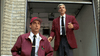 Two men in matching maroon jackets and black ties stand before an open garage; one, wearing a red cap, looks surprised as the other gestures, as if they’ve just discovered a rare Murder Most Likely (1999) DVD. Both look alert and concerned.