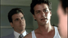 Two men indoors: one in a white tank top, serious; another in a suit and tie watching him—echoing a dramatic scene from "Murder Most Likely (1999)" DVD. The softly lit, out-of-focus background adds cinematic flair.