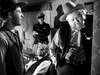 Three men in cowboy hats and vests stand in a dim room; one uses a wall phone as a police officer and an elderly man watch—a scene evoking the classic Western style of "Where Danger Lives (1950)" on DVD.
