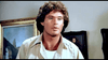 A man with curly brown hair in a light shirt stands indoors near two framed paintings, gazing off-camera with a neutral look—evoking scenes from hard-to-find movies like The Cartier Affair (1984) DVD starring Joan Collins.