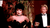 A woman in a glamorous black off-shoulder dress and sparkling necklace channels Joan Collins' elegance from The Cartier Affair (1984) DVD, as a man in a tuxedo with a drink stands behind her among decorative plants and warm lighting.