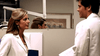 Two doctors in white coats face each other in a medical office, locked in a serious discussion reminiscent of "Too Late To Say Goodbye (2009) DVD," with the woman holding a blue folder and their reflections visible in the mirror behind them.